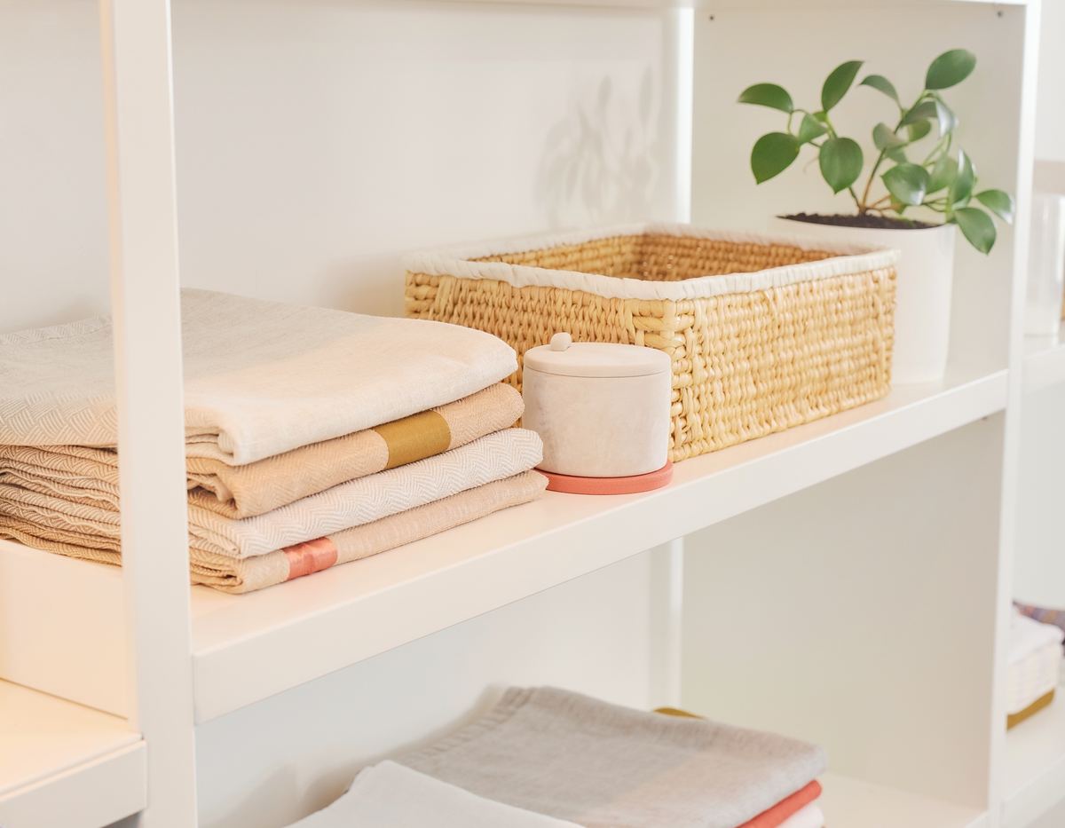 Open white shelving with neatly folded linens, a woven basket, a small lidded canister and a potted plant